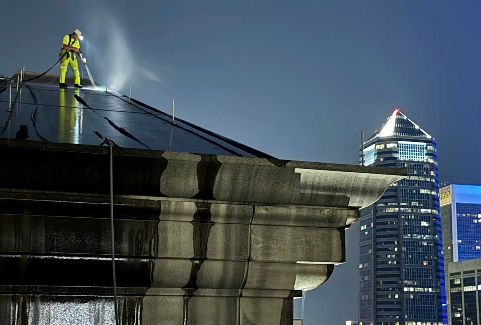 Technician pressure washing a high-rise rooftop at night above downtown Jacksonville